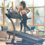 A fit woman in athletic attire running on the Nordictrack Commercial X22i treadmill. She is in a living room setting with a view of mountains, a couch, chair and tables with lamps on them
